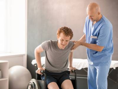 Mature clinician in uniform helping young man in pain to sit in wheelchair